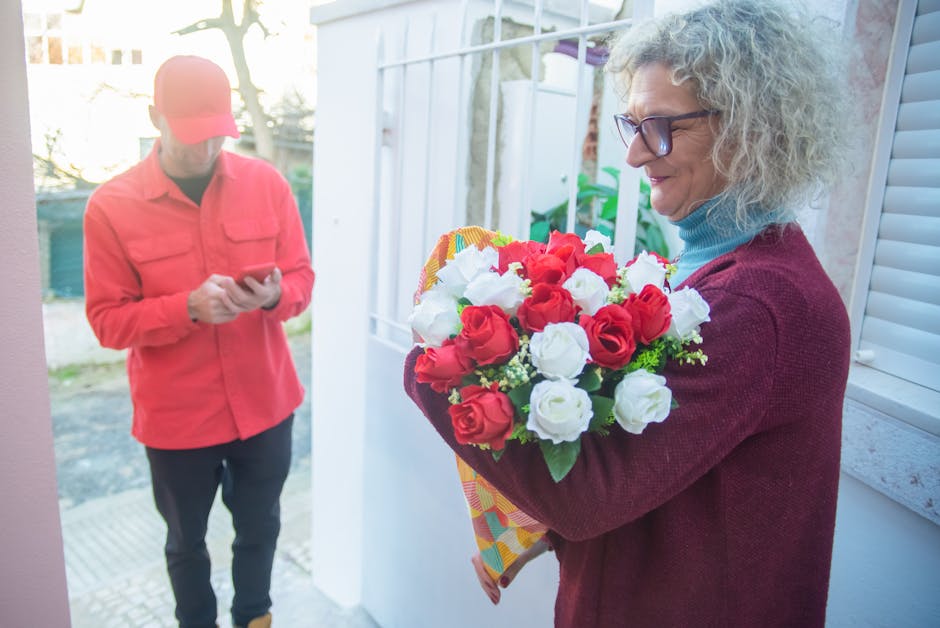 A woman with curly gray hair and glasses holding a vibrant bouquet of red and white roses wrapped in patterned paper, standing near a white door with a window. The bouquet features fresh, fully-opened
