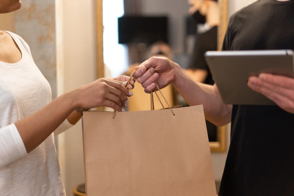 A person in a white top is receiving a floral gift, with their hand reaching out to accept a brown paper shopping bag from another individual dressed in black, who is holding a tablet in their other h