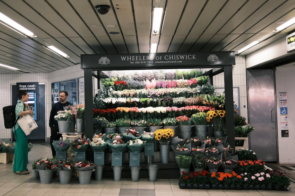 A flower stall at Finchley Central underground station, displaying an extensive variety of fresh flowers arranged in metal buckets and wrapped in teal paper. The arrangement includes roses, peonies, s
