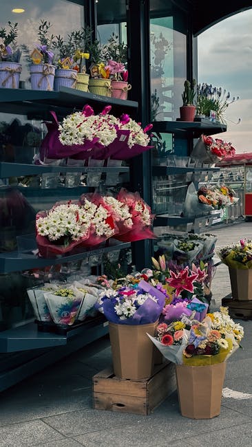 A display of various floral arrangements and bouquets outside a flower shop, including white and pink chrysanthemums, pink lilies, purple and yellow daisies, and mixed seasonal flowers wrapped in colo