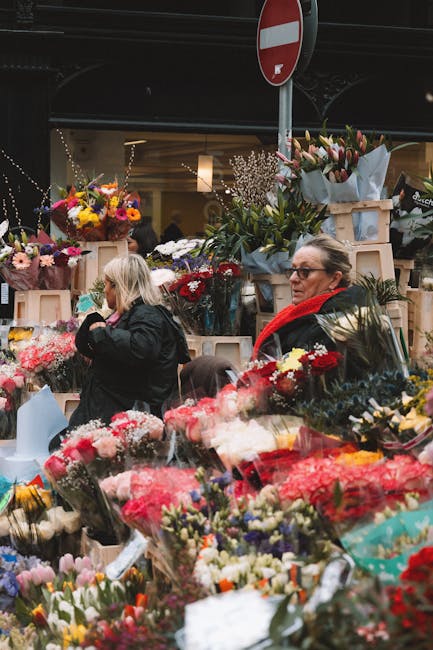 At an outdoor flower market, two women browse and purchase fresh floral arrangements from East Finchley Flowers. The scene is filled with vibrant bouquets featuring roses, lilies, tulips, and other se