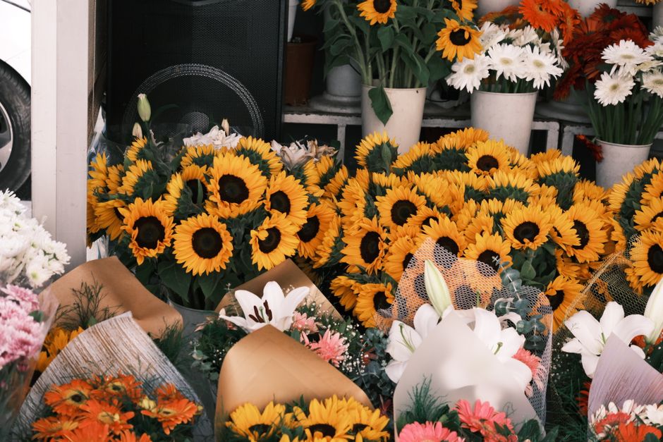 A vibrant display of fresh flower bouquets featuring primarily bright yellow sunflowers with dark centers, arranged in bundles and wrapped in brown craft paper and floral tissue. Surrounding the sunfl