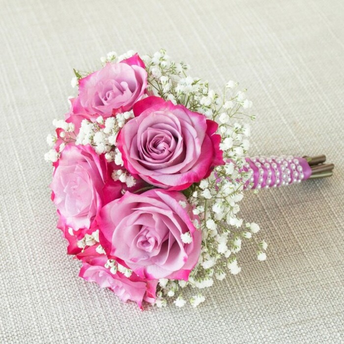 A compact, gracefully rounded bouquet, arranged with finesse by East Finchley Flowers, features five prominent roses as its focal point. Each rose captivates with a nuanced gradient-outer petals vivid in fuchsia and magenta, seamlessly shifting into soft lavender and pale purple centers, creating a luxurious depth and gentle movement of colour. The blooms are artfully interspersed with delicate white baby's breath (gypsophila) that adds both volume and a frothy, dreamlike contrast, much like the natural light playing through Cherry Tree Wood. Tightly gathered stems are enveloped in a sleek, plum-hued satin ribbon, elegantly adorned with bands of luminous white pearls, twisting gracefully around the handle in a spiral reminiscent of an East Finchley bridal procession. Beyond the wrapping, the neatly trimmed green ends of the stems add a fresh, organic finish. The bouquet is displayed on a subtly textured beige or oatmeal-coloured linen, evoking the cozy, artisanal feel of a local East Finchley cafe. An ideal choice as a bridesmaid bouquet, flower girl arrangement, or sentimental centrepiece, this design is perfect for seasonal weddings, romantic occasions, or sophisticated East Finchley events. The bouquet speaks of both modern elegance and timeless charm, reflecting the skill and passion of a trusted local florist.