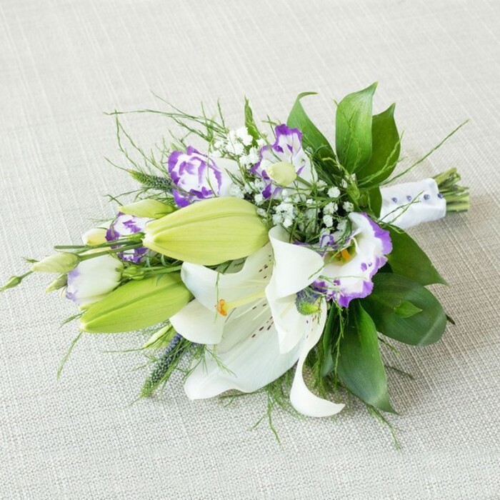 A beautifully composed, elongated bouquet lies diagonally across a subtly textured, light beige fabric background, reminiscent of a soft linen cloth spread on a table in a calm East Finchley flat. Dominating the design are several large, open white Oriental lilies, their thick, gently arching petals freshly unfurled to reveal vivid yellow-orange stamens and fine reddish-brown speckles that dot the inner surfaces. Nestled among them, multiple bright, lime-green lily buds stand upright, their smooth, tapered forms still tightly closed, adding anticipation and a sense of evolving beauty. Flowing between the lilies, ruffled Eustoma (lisianthus) flowers introduce a graceful fusion of white and purple: some blossoms are edged in delicate lavender, others in deeper violet, with cup-like blooms that appear almost painted at the margins. Fine sprays of Gypsophila are threaded throughout, their tiny white blossoms clustering like soft, suspended mist, filling the negative spaces and creating gentle visual balance. The bouquet is cradled by an abundance of greenery: broad, glossy dark green leaves lend structure and depth, while finer, wispy fronds and occasionally spikier green elements add lightness, direction, and a feeling of movement, as though touched by a breeze passing along East End Road. At the base, the gathered stems are bundled neatly and wrapped in a smooth white satin ribbon, which is subtly embellished with small pearl-like beads or dots that catch the light without overpowering the flowers. The illumination is soft and even, with no harsh shadows, allowing every curve of petal, glint of bead, and vein of foliage to be seen clearly. This bouquet carries an air of polished elegance and fresh fragrance, perfectly suited to a wedding, anniversary, or meaningful gift for someone in East Finchley, perhaps delivered from a trusted local florist for a special moment at home or in a nearby venue.