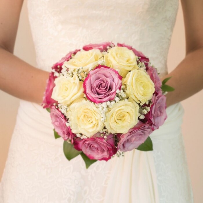 A timeless bridal bouquet unfurls in the hands of a bride from East Finchley-an image as poetic as it is precise. This close-up at eye-level accentuates the bouquet's carefully curated structure: plump, creamy lemon-yellow roses form a harmonious base, contrasted by vivid fuchsia pink and subtle lavender-hued roses that inject both romance and modern flair. Interlaced with the blooms are fine sprays of white baby's breath, each tiny flower adding ethereal softness, their delicate presence reminiscent of the gentle mornings spent in local gardens around East End Road. Hints of rich green leaves break through at the edge, their cool tones enhancing the arrangement's depth. The bride's arms cradle the bouquet just above her flowing skirt; the bodice of her white gown shimmers with complex lace embroidery-swirling patterns and leafy vines nodding to East Finchley's verdant neighbourhoods. The soft, even light imbues every petal and stitch with radiant freshness, while a blurred, neutral background guarantees the bouquet and gown remain the sole focus. This ensemble evokes devotion, artistic attention, and the uniquely personal celebrations found among East Finchley's close-knit community, offering inspiration for those seeking wedding day bouquets that capture both personality and place.