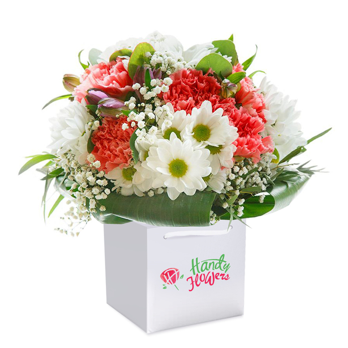 Expertly gathered into a crisp white gift bag, this abundant bouquet reflects both the refined artistry and warm welcome of East Finchley. Ruffled carnations in peach, coral, and deep red form the sumptuous core, each petal intricate and layered like the textures of Alexandra Park's flowerbeds. Snow-white daisies pop energetically alongside, their lime-splashed centers lifting the mood and lending a fresh, contemporary touch. Wispy baby's breath floats through the arrangement, softening the palette with its lacy detail and highlighting the rich, shaded greens of the foliage-a nod to the mature leafy streets winding through N2. Closed buds, brushed with purple, nestle amidst the main blooms, adding intrigue and the promise of renewal. A sturdy ribbon handle tops the square white bag, carrying the arrangement as elegantly as one might stroll down Fortis Green Road. Photographed in clean, even light, every flower radiates vibrancy and care. Whether destined for a cheerful birthday, heartfelt anniversary, or a spontaneous gesture delivered anywhere in East Finchley, the bouquet is a celebration of classic beauty and local craftsmanship, making every occasion feel special.