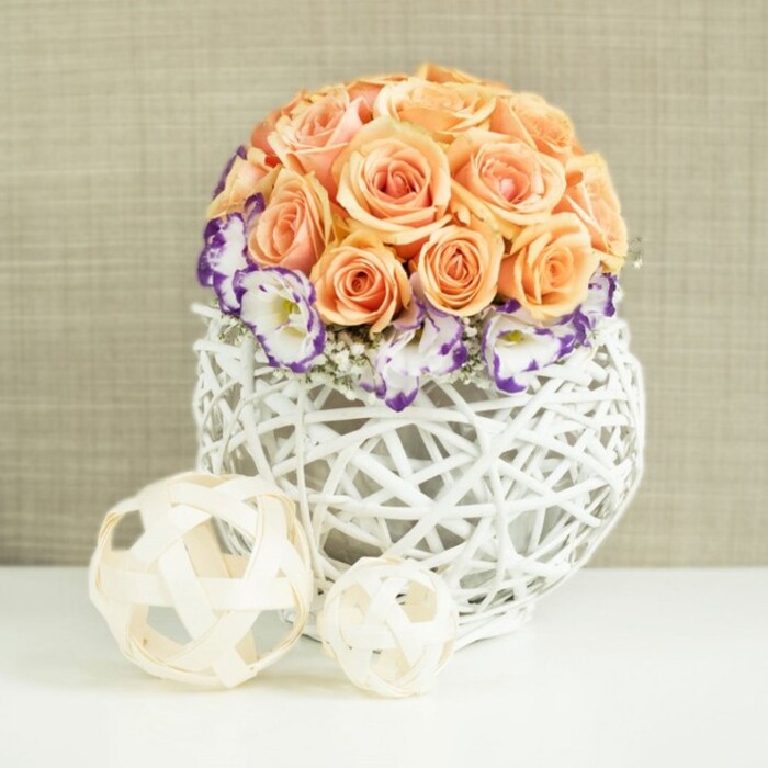A beautifully curated table arrangement is displayed on a smooth white surface, as if prepared for a wedding reception in one of East Finchley's intimate venues. The centerpiece features a perfectly rounded dome of roses, each bloom in shades of peach, apricot, and soft coral, their velvety petals unfurling fully and overlapping to form a continuous, cushioned surface. The roses create the impression of a single, sculpted form rather than individual blossoms, the colour gradations from pale blush to deeper apricot adding gentle dimension. Encircling this rose dome is a refined wreath of white lisianthus, their slightly ruffled petals edged with a strong band of deep violet or purple. This coloured rim offers a sophisticated contrast to the warm hues above. Interspersed between the lisianthus heads are fine sprigs of white gypsophila, dotting the border with airy clusters that look almost like tiny clouds gathered at the base. The flowers sit in a broad, rounded vase woven from white wicker or rattan in an open lattice pattern, the interlocking strands creating an intricate, handcrafted effect. Light filters through the gaps, casting faint shadows onto the white surface below and subtly revealing the stems within. In front of the main display, two decorative woven balls lie side by side, fashioned from narrow, natural wood strands in light beige tones; their open, airy construction echoes both the vase and the rounded form of the bouquet. Behind, a softly textured beige fabric backdrop provides a warm and neutral stage, keeping attention on the blooms while contributing to the sense of calm elegance. The overall impression is of modern romance and understated luxury, well suited to a wedding breakfast, anniversary celebration, or stylish dinner party in East Finchley, where thoughtful floral design enhances each shared moment.