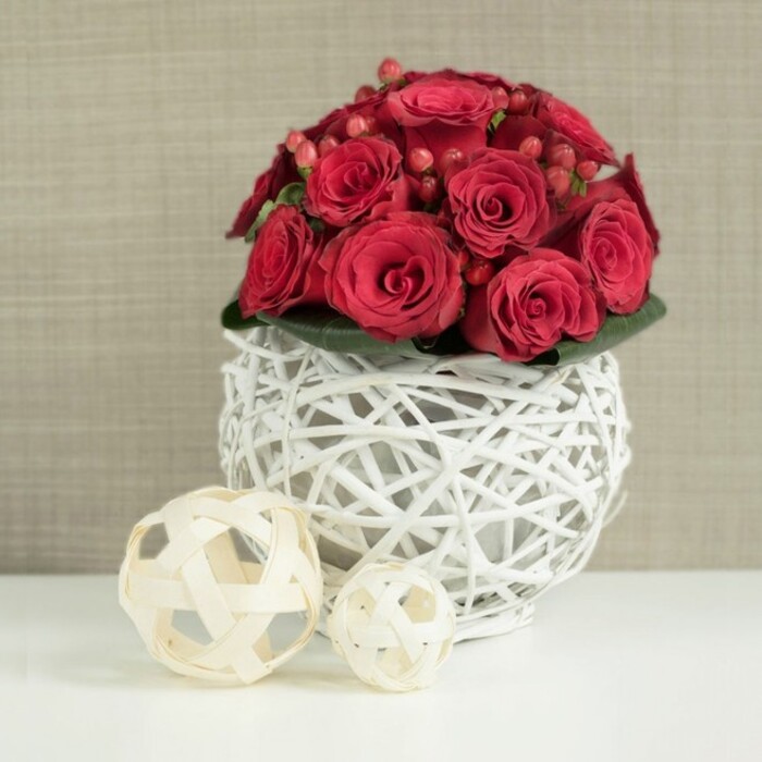 A richly coloured floral arrangement of deep red roses and berries is the clear focal point of this serene scene, resting on a bright white table that heightens every shade and texture. The roses have been arranged into a tight, dome-shaped bouquet, each head placed closely to its neighbour so that their velvety petals form a continuous, luxurious surface. Some of the blooms are wide open, showing intricate curls and gentle shadows at their centres, while others remain in more modest, half-open stages, their petals still wrapping protectively around the heart of the flower. Scattered through the mass of roses are clusters of small, glossy red Hypericum berries, which introduce a distinct, rounded texture and a slightly different tone of red, like the subtle colour changes you notice walking past the gardens near East Finchley station. At the base, a delicate ring of deep green foliage just emerges, providing a slim yet effective border that frames the lavish reds above. The bouquet is held within an unusual spherical vase formed from white-painted twigs or wicker, layered and woven into an airy orb. This openwork design lets the eye trace the criss-crossing branches, and light spills through the gaps, creating interesting shadows on the smooth white tabletop below. To the left of the main piece, two decorative balls made from pale, natural wood strips sit side by side, one slightly larger and closer to the viewer, mirroring the weave of the vase but in a softer, beige tone. The backdrop is a softly blurred wall in a light, neutral beige or taupe, reminiscent of textured plaster or fabric in a quiet East Finchley home. The composition feels romantic yet contemporary, ideal for wedding tables, anniversary dinners, or intimate celebrations in and around the High Road.