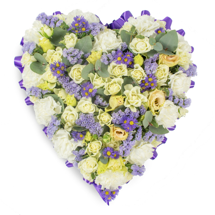 An expertly arranged heart-shaped memorial bouquet shown on a simple white studio surface, the kind of sympathetic design often chosen by neighbours in East Finchley when gathered at local chapels or commemorations. The entire perimeter is carefully edged with a deep purple satin ribbon, folded into a continuous border that gives the heart its formal silhouette and a subtle sheen that catches soft light. Within that frame, a delicate palette of whites, creams and pale yellows unfolds: clusters of cream and pale yellow roses create a plush, multi-layered texture - some buds are tightly wound, others easing open to reveal satiny interiors - while fields of white carnations add fullness with their ruffled petals and comforting presence. Interwoven throughout are sprays of lavender-coloured statice, their fine, papery clusters contributing an airy texture and a whisper of muted colour, and small violet aster-style flowers boasting sunny yellow centres that punctuate the design with brighter notes. Rounded eucalyptus leaves in a soft blue-green are placed to lift and space the blooms, offering cool tonal contrast and the suggestion of a gentle, leafy scent. The composition reads as calm and reverent, a thoughtful funeral tribute that communicates sympathy, respect and steady affection - suitable for placement at services, wakes, or graveside remembrances in the East Finchley community.