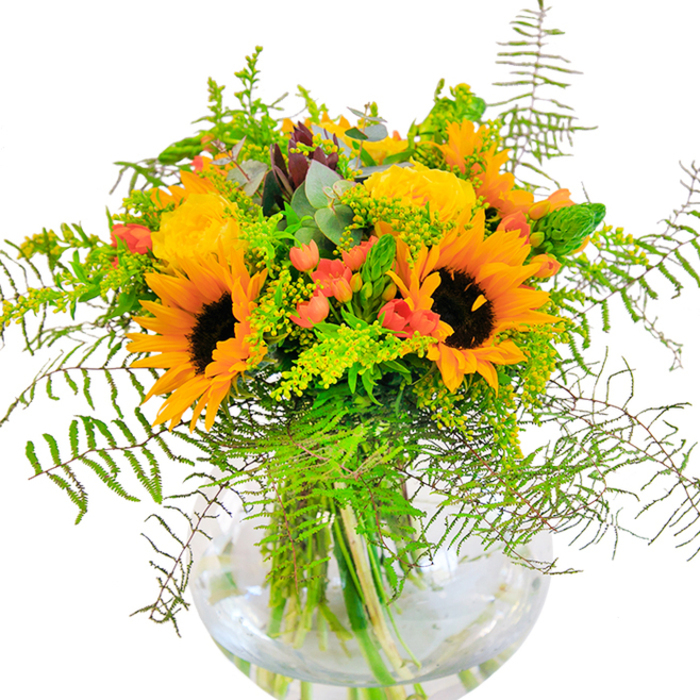 On first glance, this stunning bouquet is a burst of sunshine, expertly gathered for East Finchley homes seeking a touch of late summer's brilliance. Towering sunflowers with bold, dark centers are at the heart, their radiant yellow-orange petals unfolding with exuberance-the kind of light seen streaming through Cherry Tree Wood in early September. Entwined with the sunflowers are golden yellow garden roses, every petal curled in a classic spiral, creating an effect both opulent and timeless. Vibrant clusters of red hypericum berries pop between the rich green leaves, lending texture and a spirited, modern twist. Soft fern fronds tumble delicately from the sides, their feathery silhouettes mirroring the natural beauties along the banks of the local nature reserve. Sprays of subtle green filler, accented with miniature yellow blossoms-perhaps goldenrod or solidago-flicker through the arrangement, whispering of meadows and sunlit fields. Eucalyptus leaves, silvery and cool, peek from the bouquet, their aromatic freshness conjuring calm and sophistication. All flowers and foliage are meticulously arranged in a round, clear vase that lets the stems shine through, the water beneath sparkling with reflected afternoon light. The composition's scent mingles faintly with notes of fresh-cut greens and gentle floral sweetness, echoing the serenity found during a stroll through local parks. This arrangement, thoughtfully provided by East Finchley Flowers, is perfect for celebrating birthdays, anniversaries, or simply bringing joy to a home in the N2 postcode.