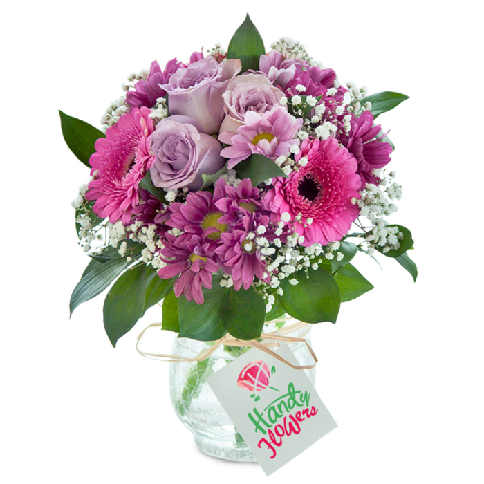 Taking pride of place before a pristine white background, this lush hand-tied bouquet celebrates the friendly charm of East Finchley. The central cluster of soft lavender roses, each bloom opening in gentle spirals, sets an aura of elegance and sentiment. Around them, bright pink Gerbera daisies-with their large radiating petals and distinct deep centers-add spirited energy, much like the vibrant buzz of East Finchley's local cafes and markets. Complementary clusters of richly colored magenta chrysanthemums with their plush petals offer inviting contrast and structure, while dainty gypsophila-fragrant and white-adds clouds of lightness, balancing the visual weight of the bolder blossoms. The lush arrangement is nestled in a clear, rounded glass vase, whose cracked texture glimmers softly in the light. A length of rustic raffia is wound with care at the neck, suspending a plain white tag suitable for messages of love or celebration. The ensemble feels at once modern and rooted in tradition, making it an inspired selection for birthdays, anniversaries, or simple gestures of appreciation throughout East Finchley and neighboring Hampstead Garden Suburb.