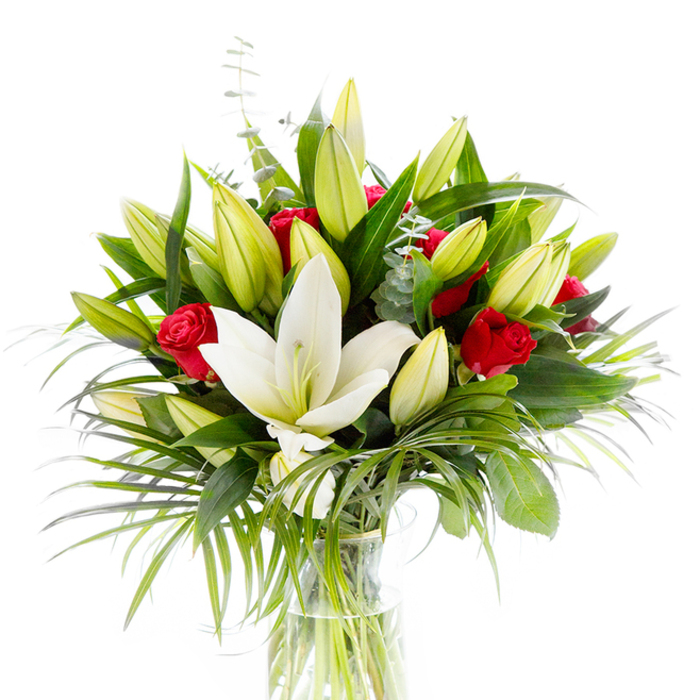 In this portrait of blooms, a single, grand creamy white Oriental lily takes centre stage, its pristine, slightly recurved petals revealing a pale yellow stamen and a tender green pistil-the serene focal point of the arrangement. Surrounding it, clusters of unopened and partially opened lily buds in lemon-lime and creamy hues create a layered rhythm that suggests growth and unfolding time. Deep, velvety red rosebuds are interspersed like small, deliberate beats of colour, their compact form promising a gradual bloom that will extend the bouquet's drama over days. The foliage is generous and varied: broad, dark forest-green leaves give solidity and depth, while lighter, arching palm-like fronds spill outward to introduce movement and air; thin stems of silvery eucalyptus with their rounded leaves soften the overall palette and add a subtle, cooling texture. The flowers sit in a simple clear glass vase, cylindrical and unadorned, where the pale green stems are visible through fresh water-an honest reveal of technique that feels both modern and classic. Shot on a bright white background, every tone and texture is clarified, from the waxy sheen of the lilies to the velvet of rose petals and the matte silver of eucalyptus. The arrangement feels appropriate for romance, milestone celebrations, or as an elegant sympathy tribute: it reads as both pure and passionate. I imagine this piece arriving with local deliveries across East Finchley-perhaps brightening a home near Creighton Avenue or a dining table by the tube-its fragrance a gentle mix of sweet lily and warm rose that invites close, quiet appreciation.