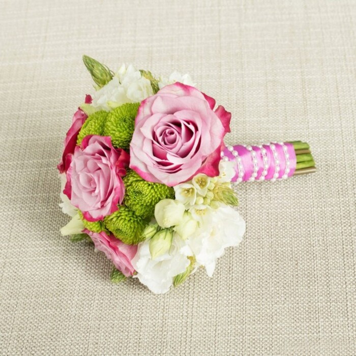 A delicately composed bridal-style bouquet rests lengthways on a softly textured, light tan fabric that recalls natural linen spread over a table in a calm East Finchley townhouse. The background's subtle weave and gentle, diffused lighting create a warm, neutral stage where every petal and pearl becomes visible. At the heart of the arrangement, three roses in full bloom command attention, especially two generous focal roses: their petals open in spirals of soft, blush pink that gradually deepen to a richer, almost magenta outline at the very edges, yielding a refined ombré effect that suggests both romance and craftsmanship. Interspersed amongst these roses, clusters of lime green button chrysanthemums form rounded, cushion-like mounds. Each pom-pom head is packed with minute florets, giving the bouquet an inviting, tactile texture and a lively shot of modern green that keeps the design from feeling too traditional. Between the larger forms, creamy white filler flowers-likely lisianthus or freesia-appear in graceful, multi-petalled sprays, lending a sense of airiness and classic bridal softness. Tiny star-shaped white flowers with sunny yellow centres glimmer in the gaps, reminiscent of waxflower, adding intricate detail that rewards closer inspection. The stems are neatly trimmed and gathered into a compact handle. This handle is wrapped in a smooth fuchsia satin ribbon, its sheen catching the light and echoing the roses' darker tones. Over the ribbon, a fine strand of white faux pearls is spiralled diagonally, like a bracelet clasped around the bouquet, introducing subtle glamour without overpowering the flowers. A short section of fresh green stems extends beyond the ribbon for a natural finish. Overall, this small but sophisticated posy feels ideal for a bridesmaid or intimate wedding at a local East Finchley venue, or as a graceful hand-tied gift delivered by East Finchley Flowers for an engagement, anniversary, or thoughtful celebration.