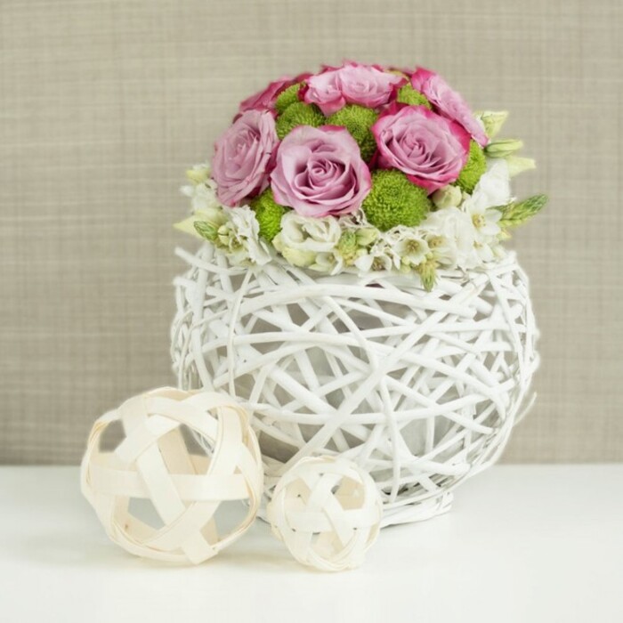 The photograph presents a lovingly styled floral composition that would feel at home on a wedding table set for guests in East Finchley, perhaps in a bright room overlooking the treetops near the High Road. At the centre, placed on a smooth white tabletop, is a unique spherical vase painted white and made from fine branches or rattan-like strips. These are intricately woven into a loose lattice, leaving open spaces that give the vase an airy, almost floating presence. Light catches along the curves and intersections of the weave, adding subtle shadows and highlights that emphasize its handmade character. Rising neatly from this base is a dome of fresh flowers arranged with precision. Lush pink and lavender roses dominate, their blooms fully open, revealing numerous velvety petals that spiral outward with a soft, romantic fullness. Some of the petals show deeper pink edges, adding dimension and visual interest to the pastel palette. Bright green button chrysanthemums are tucked throughout the roses, their tight, spherical heads standing out with a refreshing, grassy tone that enlivens the entire bouquet. At the bottom edge of the floral dome, clusters of small white filler blooms gather in a gentle ring, almost like lace around the base, easing the transition from flowers to vase and suggesting a subtle, cascading effect. To the left, on the same white surface, two decorative balls rest slightly apart, one larger and one smaller. Both are crafted from light-coloured wicker or wooden twigs woven into open, spherical forms that echo the vase's design, knitting the composition together visually. The background is a soft, defocused field of warm beige fabric, perhaps linen, with just enough texture to give depth without drawing focus from the flowers. The overall effect is serene, refined, and quietly joyful, evoking the atmosphere of a thoughtfully decorated East Finchley wedding or special occasion.