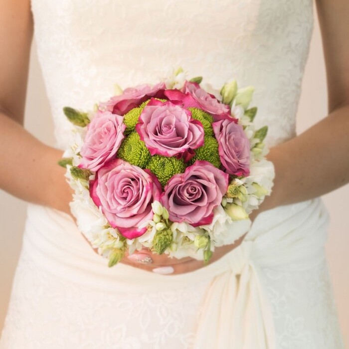 Set against the backdrop of a classic East Finchley bridal ensemble, this detailed view showcases a perfectly balanced wedding bouquet designed with both tradition and artistic flair. Central to the bouquet, mauve and dusty rose blooms unfurl in full splendor, the edges deepening to a lively fuchsia that brings a playful yet romantic accent to the round shape. Surrounding these signature roses, vibrant button mums in citrusy lime green create an enthralling contrast of texture and hue, reminiscent of spring mornings in Cherry Tree Wood. The bouquet's perimeter is encircled by clusters of petite white blooms-and hints of slender, vivid green foliage-contributing to an elevated sense of freshness and formality. The bride's hands, carefully manicured in pale blush and finished with a single sparkling highlight, cradle the bouquet with gentle reverence, her dress reflecting the light in elegant swathes of ivory lace and understated drapery. The softly diffused natural lighting draws out the intricacy of the floral arrangement and the delicate patterns of the gown-a hallmark of artistry celebrated by local East Finchley florists. This composition, ideal for wedding floral inspiration or explorations of colour palettes, radiates a tranquil yet joyful spirit that defines memorable North London ceremonies.