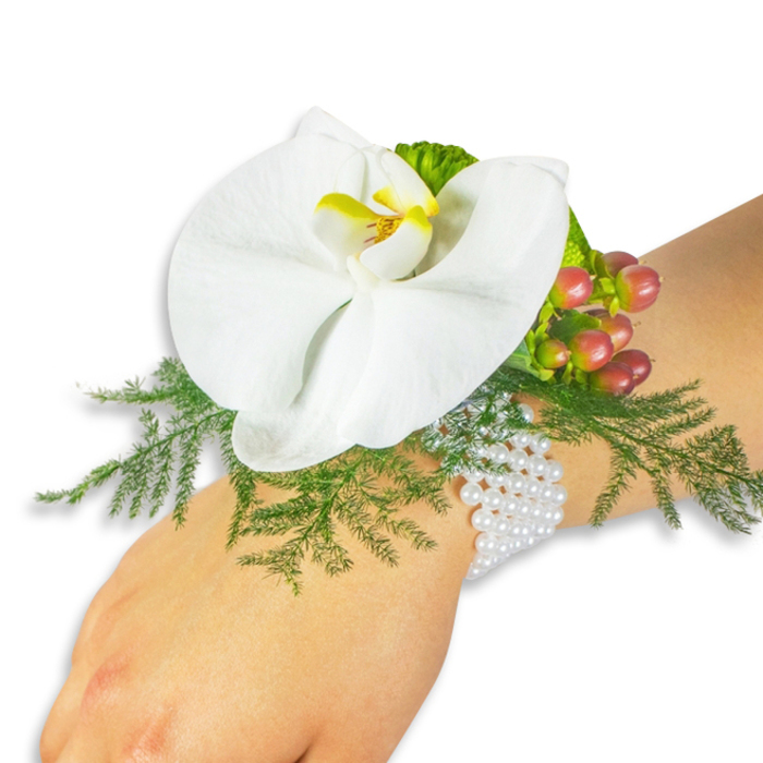 Presented on a light-skinned wrist against a pristine white field, this elegant wrist corsage reads as a study in contrast and tactile pleasure. The centrepiece is a large, immaculate white Phalaenopsis orchid whose broad petals unfold in gentle curves; the orchid's throat shows a concentrated yellow centre flushed with a deeper orange, a warm focal point that draws the eye. A generous bed of asparagus fern-bright, feathery and finely textured-spreads beneath the bloom, its filigree giving the piece an airy, botanical silhouette. Nestled to the upper right, a compact cluster of Hypericum berries offers seasonal colour, the individual berries graduating from pale green to soft rose and finally to a deep cranberry hue, each glossy orb catching the light like tiny polished stones. Lime-green, slightly ruffled accent foliage peeks through to add freshness and dimensional contrast. The arrangement is secured by a classic multi-strand bracelet of small white pearls; their satiny sheen and slight heft lend an heirloom quality while the flexible construction keeps the corsage comfortable for long wear. Soft, even lighting highlights the orchid's satiny surface, the fern's delicate veins and the berries' reflective skins, producing a calm, intimate mood that suggests quiet celebration-perfect for a wedding, graduation, or prom. The craftsmanship suggests a local florist's careful hand, a piece that would sit beautifully on wrists across East Finchley and nearby Fortis Green for those who favour understated, timeless floral accessories.