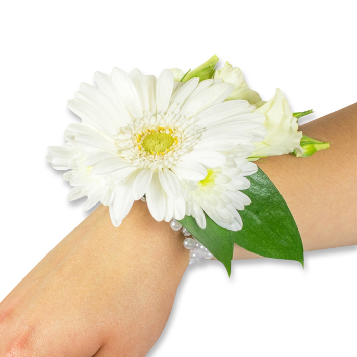 A striking white wrist corsage is displayed across a fair to medium forearm, captured against a spotless white background that accentuates every petal and glossy leaf. The dominant bloom is a large white Gerbera daisy, its clean, symmetrical petals spreading outward from a textured, yellow-green centre that reads almost like a tiny pincushion and offers a fresh, uplifting focal point. Wrapped closely around this centerpiece are clusters of small white chrysanthemums or spray mums and several creamy lisianthus - some in tight bud, others beginning to unfurl - which together create a layered, romantic texture and soft visual rhythm. The floral mass is given weight and contrast by rich, deep green salal or ruscus leaves, their broad, reflective surfaces tucking behind and beneath the blossoms to frame them and extend gently along the wrist. The corsage is secured by an elegant, multi-strand white pearl bracelet that glows softly under the light, lending the design a classic refinement suitable for brides, bridesmaids, prom attendees, and family members at graduations or anniversaries. The scent is quietly green and floral, the petals offering a cool, almost watery softness if brushed by fingertips. As a local florist in East Finchley, we often assemble pieces like this for couples walking through Cherry Tree Wood to their ceremony, or for families gathering near the Market Place; East Finchley Flowers crafts each corsage to sit comfortably through photographs and lengthy celebrations, balancing freshness, proportion, and long-lasting bloom quality. The photograph's crisp lighting reveals the fine details of petal edges and beadwork, underscoring the careful handcraft that turns fresh stems into an elegant, wearable keepsake.