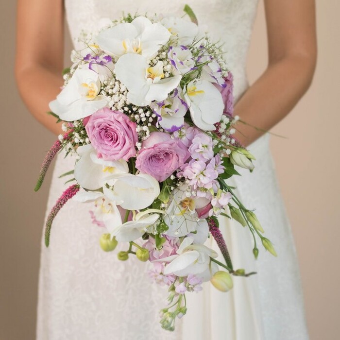Rich in texture and colour, this visual narrative unveils a bride in East Finchley clasping a breathtaking waterfall bouquet-the product of expert hands and local passion. Prominent, broad Phalaenopsis orchids in crisp white shimmer in the soft ambient light, their golden-toned centres reflecting the understated luxury of a handpicked arrangement. Encircling the orchids, you'll find layers of blush pink and pale lavender roses, their velvety petals opening generous hearts that evoke romance and the natural beauty of North London's green spaces. Gentle sprays of gypsophila, with each tiny white blossom floating amidst the larger flowers, bring a sense of airiness and movement. At the trailing edge of the bouquet, deep magenta Amaranthus spills downward, its distinctive, fluffy texture and saturated hue painting a dramatic line through the composition, a flourish that feels right at home in East Finchley's creative spirit. Interspersed are accents of soft purple blooms-perhaps sweet peas or stock-adding complexity and subtle scent to the ensemble. Sprigs of fresh green leaves and slender buds peek between blooms, grounding the arrangement with natural freshness. The bride's dress, awash in white and accented by refined lace, sets an elegant stage for the bouquet's lively palette. Altogether, this bouquet is a harmonious blend of opulent whites, delicate pinks, serene lavenders, and striking magenta-a tribute to love, artistry, and the vibrant local culture of East Finchley, ready to grace any memorable North London wedding.