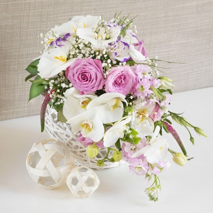 A refined floral arrangement in a white wicker basket is shown in a soft, close-up composition, echoing the understated elegance often seen at East Finchley weddings and special gatherings. The basket is filled with a generous mix of pastel pink and pure white blooms, forming a rounded yet slightly free-flowing display. At the centre, large pink roses open fully, each one shaped into a near-perfect spiral with velvety petals fanning outwards in layers, providing a romantic focal point. Graceful white Phalaenopsis orchids curve through and above the roses, their wide, rounded petals and distinctive yellow-orange centres lending a sophisticated, contemporary bridal touch. Among them, white lisianthus blooms appear with frilled petals, a few edged in cool lavender, creating subtle shifts in tone that add visual interest without overpowering the gentle colour scheme. Pale pink stock flowers rise and then bend slightly over the basket's edge, their many small florets clustered along each stem suggesting light fragrance and adding a soft, airy movement, as though stirred by a breeze from nearby Cherry Tree Wood. Fine stems of baby's breath lace through the entire composition, forming delicate clouds of tiny white flowers that fill gaps and give the arrangement a gossamer-light feel. Interspersed greenery, mostly slender leaves and tender buds, peeks between blossoms and trails minimally from the sides, anchoring the bouquet in a natural, garden-inspired aesthetic. The white wicker basket, with closely interwoven strands and a smooth painted finish, sits on a bright white surface next to two decorative spheres crafted from pale, twisted twigs, their open lattice design echoing the basket's texture and adding rustic charm. Behind, a neutral, beige-toned fabric forms a soft, textured backdrop, ensuring the colours and shapes of the flowers remain the focus. This arrangement would feel perfectly at home as a wedding table centrepiece or a romantic gift for an East Finchley home.