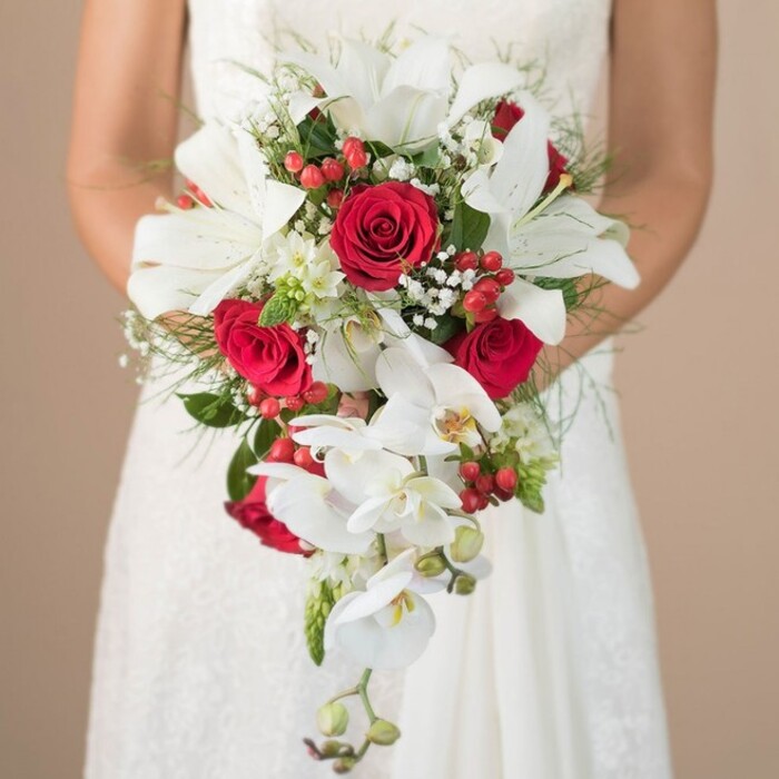 This radiant photograph spotlights a bride in East Finchley, waist-up, wearing a flowing, softly textured white gown with delicate lace details. She gently holds a truly exquisite cascading bouquet, its design a harmonious fusion of tradition and modern style. The focal point is a cluster of dazzling white lilies-notable for their crisp, trumpet-shaped blooms and aromatic allure-accented by abundant white phalaenopsis orchids with trailing sprays that bring fluidity and grace. Crimson red roses are nested throughout, their velvety petals unfurling to reveal a richness of tone and a sense of passionate energy, reminiscent of romantic strolls along the quiet avenues of East Finchley. Tiny sprigs of baby's breath and bursts of glossy hypericum berries offer a playful texture and dynamic color contrast, dotting the bouquet with touches of celebration. Wispy green asparagus fern and broad, deep-hued leaves provide subtle architectural support and a fresh vibrancy akin to the greenery of Coldfall Wood. The bride's bare arms embrace the bouquet with gentle poise, all set against a creamy, neutral background that ensures each floral nuance stands out. This bouquet, expertly arranged for a classic North London ceremony, is the embodiment of romance, artistry, and the understated sophistication that defines East Finchley.