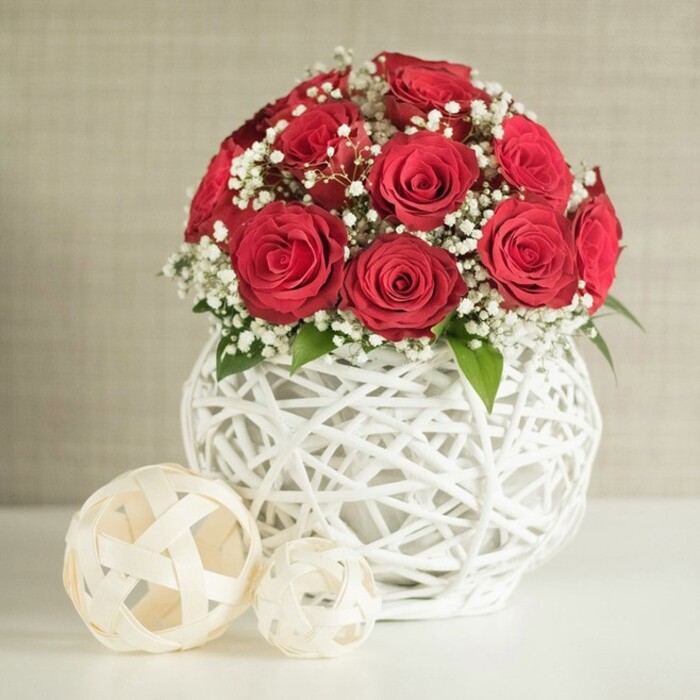 A romantic table centrepiece of crimson roses and delicate gypsophila is presented against a calming neutral backdrop, evoking the refined yet welcoming atmosphere of an East Finchley celebration. At the heart of the composition is a generous dome of deep red roses, bunched closely together to form a rich, full mound of velvety petals. Each flower is fully open, showing off its intricate spiral of petals that curl outward in soft layers, creating depth and a sense of luxurious abundance. Between the roses, clusters of white baby's breath drift gently, their minuscule blooms forming soft, cloud-like patches that lighten the arrangement and add a hint of airy whimsy. Small accents of fresh green foliage appear where stems meet blooms, introducing a quiet touch of nature that suggests the cool, leafy streets around N2 on a spring morning. The bouquet is gathered in a distinctive round white vase, its surface crafted entirely from interwoven white strands that criss-cross to form an open lattice. This woven structure allows the underlying wall colour to peek through and casts delicate shadows, giving the vase a sculptural, artisan-made quality that would feel at home in a stylish flat near East End Road. Resting on a smooth, pale tabletop, the vase is complemented by two decorative woven balls positioned slightly to the left. These off-white or cream spheres, one larger and one smaller, share the same relaxed, open-weave character, tying the decor together and reinforcing the handcrafted feel. Behind, a softly textured wall in a light beige or warm stone hue recedes gently out of focus, providing a serene canvas that ensures the deep red roses remain the clear focal point. The result is an arrangement that feels intimate, graceful, and perfectly suited for wedding tables, anniversary gatherings, or romantic dinners across East Finchley.