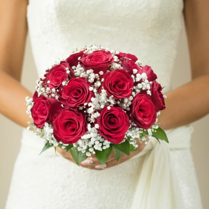 In the heart of East Finchley, this stunning bridal portrait brings the beauty of classic wedding florals to life. The focal point is a domed bouquet, brimming with rich, scarlet red roses at various stages of bloom-their silky, well-formed petals lending the display a sensual depth reminiscent of vibrant spring gardens near Cherry Tree Wood. Weaving through these passionate reds are gentle clusters of baby's breath, their petite, pristine white blossoms softening the bouquet and evoking a sense of purity and cloud-like grace. At the ensemble's base, a sprinkle of verdant green leaves shines through, offering a lively nod to the neighbourhood's leafy avenues and parks. The bride's tender hold, adorned with manicured nails, secures the bouquet just above the crisp, delicately textured bodice of her wedding dress, which may feature local embroidery or lace traditions. A soft, trailing sash elegantly falls to one side, completing her look. The background is blurred in creamy, neutral hues, ensuring that every floral and fabric detail stands out. The image beautifully reflects what East Finchley Flowers is known for: expert craftsmanship, wedding-day romance, and a deep connection to the tastes and atmosphere of the N2 community, making it a perfect reference for couples dreaming of an elegant, memorable celebration.