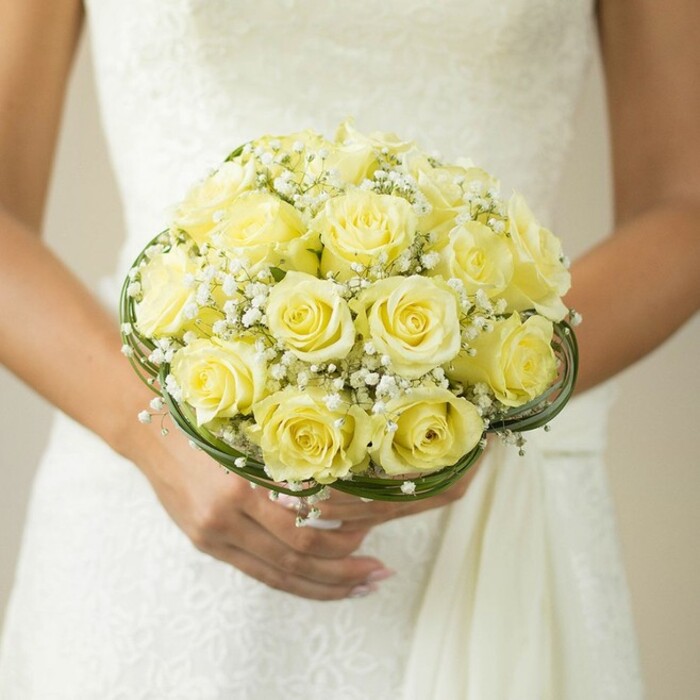 A radiant bridal bouquet is lovingly displayed in the heart of East Finchley, caught in an intimate close-up that renders each detail with tactile clarity. Soft, pale yellow roses-dozens of them-form a densely clustered, rounded shape, their petals gently unfurling in a harmonious blend that reflects the expertise of local florists crafting for special moments. Interlaced throughout the arrangement, dainty sprays of baby's breath impart an ethereal lightness, visually softening the yellows just as a gentle early afternoon breeze might drift through the parks by Fortis Green. At the base, elegant loops of dark green foliage or slender decorative wire outline the bouquet, providing a sophisticated foundation reminiscent of custom floral work found only at East Finchley Flowers. The wearer's hands-manicured with pale pink polish, a glinting ring on the left hand-hold the bouquet tenderly, emphasizing the significance of the moment. In the background, the textural nuances of a white wedding gown-potentially lace or embroidered-hint at personal and local tradition. The softly blurred setting maintains an intimate and romantic atmosphere, highlighting the artistry and community spirit that define wedding floral creations in this North London neighbourhood.