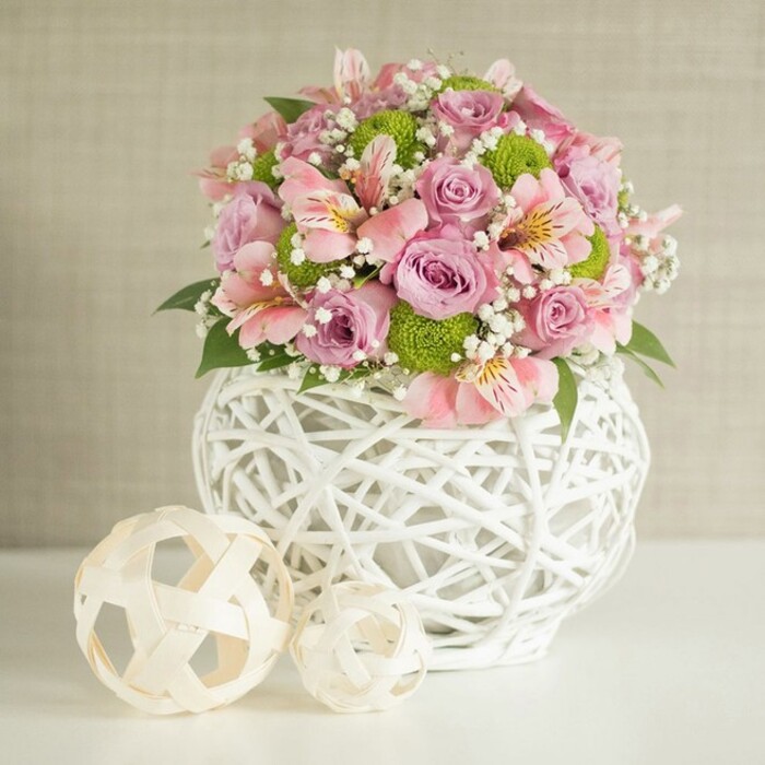 A spectacular wedding centerpiece sits gracefully atop a white table, illuminated by soft, diffused daylight, echoing the relaxed, welcoming mood of East Finchley's leafy streets. At its core, layers of lush pastel pink and lavender roses unravel with velvety petal softness, their beauty enhanced by slender pink alstroemeria blooms marked with distinctive streaks, and lively bursts of lime green button chrysanthemums. Baby's breath is scattered throughout in delicate white clusters, adding an ethereal, cloud-like quality, while rich emerald leaves emerge here and there, lending the arrangement depth and liveliness. The flowers are arranged in a striking white vase-that distinctive open-lattice design, sculpted from fine woven strips, is both contemporary and nostalgic, reminiscent of art deco details found around Tally Ho Corner. On the table beside the vase, two decorative spheres in neutral, natural hues are carefully positioned: the larger orb sits closer and echoes the vase's open-weave pattern, while its smaller companion brings a playful, artistic finish. The subtly textured light beige background ensures that each nuance of colour and texture stands out, from the creamy pinks to the vivid greens. This arrangement's gentle sophistication and thoughtfully layered textures make it an ideal centrepiece for weddings, milestone celebrations, or East Finchley gatherings where flowers speak of both beauty and place-a true reflection of the neighbourhood's community spirit and attention to craft.