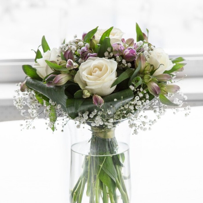 A beautifully balanced bouquet of white roses, lavender alstroemeria, and delicate baby's breath sits gracefully in a straight-sided clear glass vase, bathed in soft natural light that feels like it could be streaming in from a bay window on a calm morning in East Finchley. The roses, a creamy, almost ivory white, form the central focus: each bloom fully open yet still fresh, with layered petals that curl and fold like finely pressed satin, creating a plush, romantic texture. Nestled around them are clusters of alstroemeria in muted shades of lilac and dusty purple, some flowers unfurled to reveal their graceful, lily-like form, others still held in elongated buds tinged with green sepals, giving the arrangement a sense of gentle movement and unfolding life. Scattered between these larger flowers, tiny star-like blossoms of baby's breath float in airy sprays, lending a cloud-soft, almost sparkling lightness that fills in the gaps without ever feeling crowded. Lush greenery frames the ensemble: generous dark green leaves and more slender, arching stems cradle the blooms, adding structure, contrast, and a feeling of garden freshness, as if the stems might have been cut that morning from a well-tended plot near Cherry Tree Wood. The clear vase reveals the stems below the waterline, carefully trimmed and crossed in a deliberate, almost architectural pattern that reflects the florist's skill and attention to detail. The overall mood is peaceful and hopeful, a harmonious blend of white and soft purple that would be equally at home as a sympathy tribute in a quiet living room off East End Road, as a graceful anniversary gift delivered to a couple in nearby Muswell Hill, or as an elegant centerpiece for a small gathering where warmth, reflection, and understated beauty are cherished.