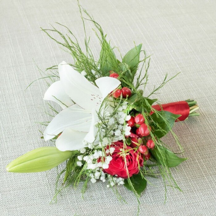 A charming, thoughtfully crafted bouquet for a bridesmaid is showcased against a softly textured, light linen fabric-a scene that recalls the calming, elegant interiors synonymous with East Finchley residences. Anchored by a radiant white lily at the centre, each petal spreads outward with tranquil grace, delicately adorned with subtle reddish-brown spots near the golden anthers, capturing both freshness and poise. To the lower right, a velvety rich red rose offers its glorious spiral of petals-a timeless statement of love that draws inspiration from the local history and storybook gardens found in East Finchley. Scattered among the main flowers are dainty tufts of white gypsophila, bestowing a sense of weightless brightness. Bold red berries, glossy and inviting, nestle artfully between blossoms, evoking memories of seasonal markets along East End Road. On the left, a slender green lily bud hints at future blooms and unspoken hopes. Surrounding greenery, ranging from fine, fernlike fronds to robust, deep-hued leaves, creates a textured, botanical canopy. The entire bouquet is pulled together with a sleek, red satin ribbon at the stems, harmonising with the reds above. Radiating a mood of celebration and delicate romance, this bouquet is meticulously designed for special occasions-weddings, anniversaries, or heartfelt moments-in and around East Finchley, reflecting both artistry and a sense of local tradition.