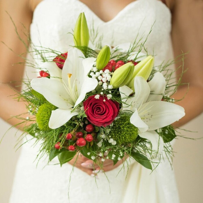 The lens lingers on a moment of East Finchley wedding bliss, drawing attention to the bride's softly clasped hands and the breathtaking bouquet she carries. The graceful upper line of her white, delicately textured gown sets the stage for an arrangement that is as thoughtful as it is vibrant. Pristine oriental lilies flare open at the heart of the bouquet, their elegant petals and golden centres glowing with purity-fitting for ceremonies near the tree-lined avenues of N2. Deep red roses, their velvety swirls tightly coiled, sit amid the lilies, conveying enduring passion and classic romance against the creamy backdrop. Petite, fresh green lily buds are scattered throughout, bringing a hint of lively anticipation. Wispy clouds of baby's breath infuse an ethereal air, while splashes of red from hypericum berries and gentle green spherical blooms evoke playful whimsy, mirroring the area's lush green lanes. Anchoring the bouquet is a lush assembly of foliage in diverse shapes and tones, echoing the hand-in-hand vibrancy of East Finchley life. The composition, framed by the bride's careful embrace and a softly blurred background, is at once elegant and exuberant, making it a natural choice for celebrations styled by East Finchley Flowers, or as inspiration for local wedding planning and floral artistry.