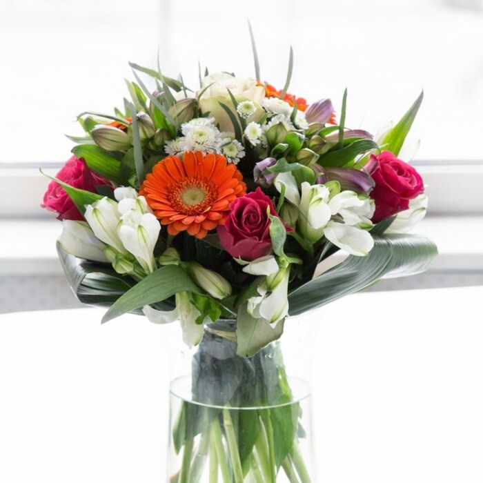 This exquisite bouquet, handcrafted for the East Finchley community, seamlessly blends radiant colours, sophisticated structure, and a sense of heartfelt emotion. The arrangement bursts into view with bold orange and soft peach gerbera daisies, their luminous petals wrapping around deep brown eyes in a spirited embrace that reflects the welcoming nature of East Finchley's neighbours. Nestled nearby, spiralling magenta roses bring depth and elegance, their plush whorls unfolding like treasured secrets. Among these lie clusters of multi-petaled white alstroemeria and compact spray roses, each one casting a delicate shimmer and brightening the ensemble with grace. Soft pink accents and wispy white fillers emerge from within, layering the bouquet with subtle richness. The entire composition is held together by artfully curved aspidistra leaves-a verdant, glossy collar that grounds the exuberant florals and frames the arrangement with a sense of refinement. Additional pointed leaves and airy sprigs fill each interval, lending both volume and harmony. The tall, clear glass vase allows the bouquet's sturdy green stems to be showcased in a way that recalls a fresh harvest from one of East Finchley's secluded gardens. Gentle ambient light suffuses the scene, enriching the natural vibrancy of every hue. This creation would be perfectly suited to a party on The High Road, a heartfelt reunion, or simply making someone's day on Twyford Avenue. Each element is expertly chosen and arranged, reflecting the care, craftsmanship, and community pride of a dedicated East Finchley florist.