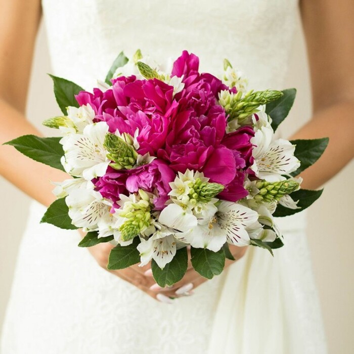 The image shows a close, intimate view of a bride holding a hand-tied bridal bouquet, pressed gently against the bodice of her white lace gown so that the intricate floral pattern of the dress forms a quiet backdrop to the vivid colours. Dominating the centre of the bouquet are full, deeply ruffled peonies in shades of fuchsia and magenta, their many petals overlapping in rich, rounded layers that convey both luxury and softness. Threaded through these larger blooms are stems of white alstroemeria, also known as Peruvian lilies, each flower opening into a star-like shape with inner petals marked by fine, maroon speckles and subtle striping that draws the eye inward. Around and between the main blossoms, slender green, spiky buds and small starry filler flowers reach upward, adding texture and a fresh, just-picked feeling, as though the stems might have come from a morning delivery to a florist on East Finchley High Road. The entire composition is encircled by broad, deep green leaves with a slight sheen, creating a natural frame that enhances the contrast between the hot pink tones and the crisp white petals. The bride's hands are visible at the base of the bouquet, fingers curled gently around the bound stems, her nails painted a delicate light pink that echoes the romantic palette of the arrangement. Soft, diffused light bathes the flowers, bringing out the velvety quality of the peonies and making the whites appear luminous against the dress, while the background falls into a gentle blur so that nothing distracts from the bouquet and the bride's midsection. The effect is one of joyful elegance, perfect for a spring or summer wedding in East Finchley, where couples might choose a bouquet like this to bring a burst of colour and fragrance to their ceremony and photographs.