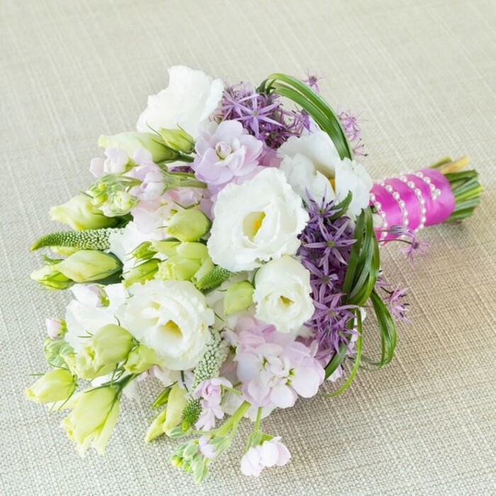 Arranged with a local florist's mastery, this radiant bouquet is laid on a softly woven beige linen, reminiscent of the artisan textiles found in East Finchley's boutique shops. Large, fully open Lisianthus blooms in creamy white radiate a subtle, peaceful warmth-central to the bouquet's harmonious appeal-while elongated green buds hint at evolution and gentle optimism. Threaded amid the whites are plumes of stock in shades of lavender-pink, conjuring a soft, airy mood echoing the neighbourhood's spring mornings. Deep violet alliums, their spiky star-bursts seeking attention, add structure and daring contrast-reflecting perhaps the creative spirit thriving in East Finchley. Swathes of vivid bear grass encircle the main blossoms, their arcs drawing the eye and suggesting the rhythm of nearby parks. Dotted green fillers and tiny buds interlink the composition, offering texture and layers. The stems are tightly wrapped in a bold magenta satin, crisscrossed elegantly with pearly accents that glint softly under the natural light commonly found streaming through Muswell Hill windows. This bouquet, perfect for a sunlit wedding or stylish party at a community hall, captures the heart of East Finchley-driven by local artistry, fresh seasonal flowers, and a palette that is both timeless and on-trend.