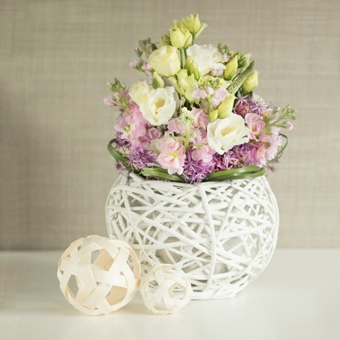 Carefully composed with an eye for harmony, this table arrangement weaves together the soft pastel palette and organic textures favoured by East Finchley's discerning residents. Central to the display, plentiful stems of creamy eustoma and gentle yellow lisianthus curl with lush ruffles next to vertical notes of pale pink stock, their aroma a subtle nod to English cottage gardens. Beneath, rich violet filler flowers form a pillowy base, their tiny blooms enhancing visual weight and interest, while a wreath of glossy green foliage completes the bouquet's inviting form. These botanicals are gathered in a modern open-weave vase, meticulously shaped from painted white rattan, its spherical silhouette echoing both contemporary style and the timeless artistry of local craftspeople. To the left of the vase, a pair of pale wooden spheres-one slightly larger, each with a lattice-like design-echo the arrangement's themes of nature and structure, much like the sculptural details spotted in East Finchley's gallery shops. The arrangement sits atop a luminous white surface, its background a warm greige, lightly textured, reminiscent of a Finchley sitting room filled with morning sunlight. This image encapsulates the spirit of celebrations held in local venues or family homes, delivering not just blooms but the fresh, handcrafted elegance that East Finchley Flowers has become known for.