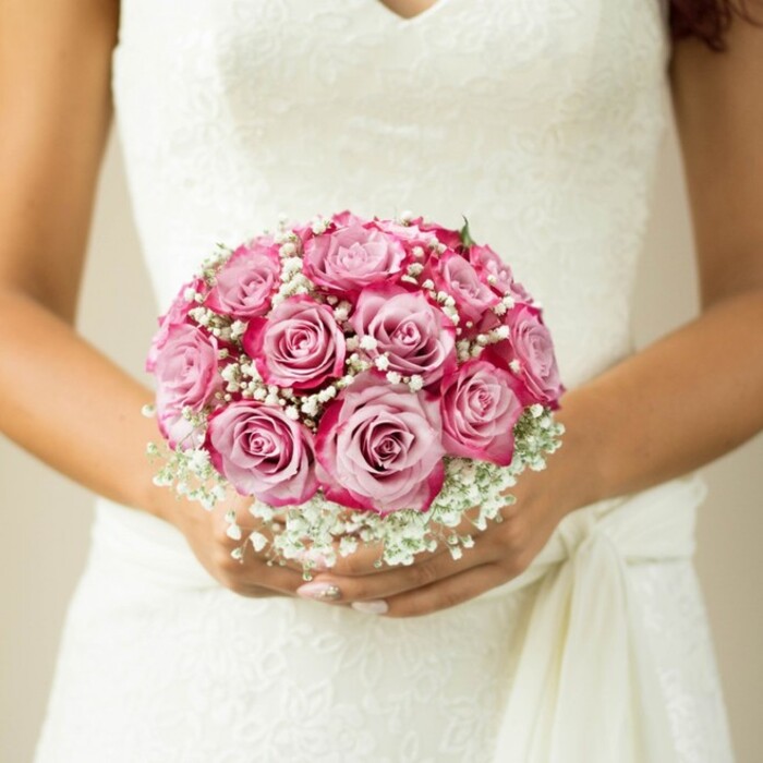 This tender close-up depicts a bride in East Finchley, arms delicately encircling a hand-crafted, round bridal bouquet that radiates sophistication and care. The composition is lush with medium-sized roses, each petal unfurling with a gradient from gentle blush to vibrant fuchsia-a natural progression reflecting the romantic landscapes of Hampstead Garden Suburb. Among the roses, trailing sprigs of feathery white gypsophila lend a soft, billowy texture that drapes the arrangement in a dreamlike glow, reminiscent of wedding mornings at Alexandra Palace. The bride's hands, just visible beneath the cluster of blooms, are finished with light, pale polish that heightens the understated luxury. Her sleeveless dress features an opulently embroidered lace bodice and a folded sash detail on the right, the fabric gathered gracefully as seen in classic North London ceremonies. The backdrop-a muted wash of beige-is softly blurred, letting the vivid artistry of the flowers and the bride's gentle posture shine through. Overall, this image radiates the elegance and heartfelt significance of a locally designed bouquet, capturing the timeless beauty and emotion found in East Finchley's classic wedding scenes.
