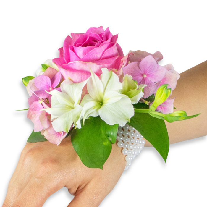 On a stark white background, a refined floral wristlet is displayed on the left hand and forearm of a person with a light-to-medium skin tone, the composition capturing both floral detail and jewellery-like craftsmanship. At the heart of the corsage sits a striking deep fuchsia-pink rose, its heavy, layered petals forming a rich, velvety centre that draws the eye. Surrounding blooms of soft blush-pink hydrangea create a plush, textured cushion that softens the silhouette and adds volume. Interspersed throughout are crisp white alstroemeria (Peruvian lilies), their four to six petals edged with a pale lime-green trace at the throat, lending a delicate, refined contrast. To the right, a small cluster of pale green alstroemeria buds and a few tiny pink buds offer a promise of unfolding petals, adding a natural, living rhythm to the arrangement. Several broad, glossy emerald leaves cradle and lift the flowers, their deep green tones intensifying the brighter hues. The corsage is fastened with an elegant multi-strand bracelet of tiny white pearls, which encircle the wrist and extend slightly over the back of the hand; the pearls' satiny sheen and tactile smoothness complement the soft textures of the blooms. Gentle, diffused lighting highlights petal margins and leaf veins, giving the piece a polished studio look without harsh contrast. Crafted with local care, this design is representative of our East Finchley aesthetic and is available for timely delivery across East Finchley and neighbouring Highgate and Muswell Hill through East Finchley Flowers, making it an ideal wearable accent for weddings, proms, or intimate celebrations.