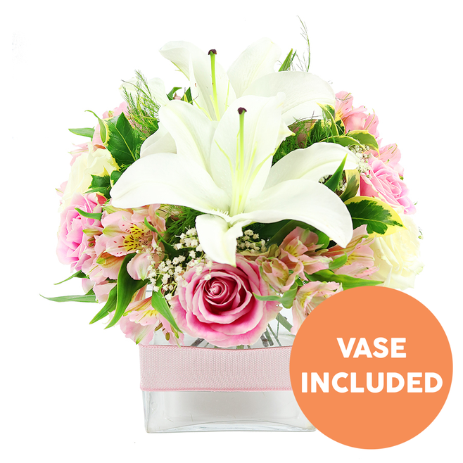 This image shows a beautifully balanced bouquet of white and pink flowers set against a pristine white background, much like the bright interiors of East Finchley apartments near Muswell Hill. At the heart of the arrangement, two impressive white Oriental lilies sit in full bloom, their large, trumpet-like petals slightly recurved and satin-smooth, cradling prominent dark reddish-brown stamens that stand upright from the center. Surrounding them is a generous collar of soft pink roses, some still in tight bud, resembling perfect little spirals, while others have opened more fully, their velvety petals layered in gentle ruffles that speak of affection and romance. Among these focal blooms, stems of alstroemeria thread their way through, bringing notes of pale pink, blush, and soft peach, with inner petals delicately freckled with yellow and fine streaks that add visual interest and texture. Fine clusters of white baby's breath sit lightly in the gaps like tufts of cloud, giving the entire design an airy, open quality. The arrangement is framed by rich green foliage in several forms: broad, glossy leaves reflect the light, while finer, feathery greens give a sense of lightness and movement, echoing the leafy character of nearby East Finchley streets. All the stems are neatly arranged in a low, square clear glass vase that provides a clean, modern base without competing for attention. Around the vase's midsection, a pale pink satin ribbon is wrapped in a horizontal band, subtly enhancing the romantic tone and hinting at a special occasion. In the bottom right corner of the image, a circular orange label with bold white text reads VASE INCLUDED, clearly highlighting that the glass container comes with the flowers. The overall feel is fresh, elegant, and thoughtful, ideal for anniversaries, birthdays, or simply adding a note of beauty to an East Finchley home.